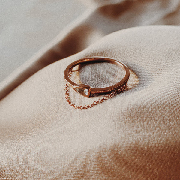  Rose gold ring with bezeled pear-shaped gem with dangled chain.